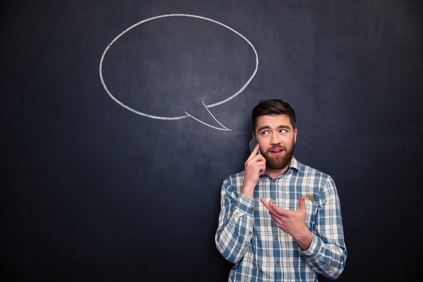 Happy handsome young man with beard talking on mobile phone standing over chalkboard background with blank speech bubble