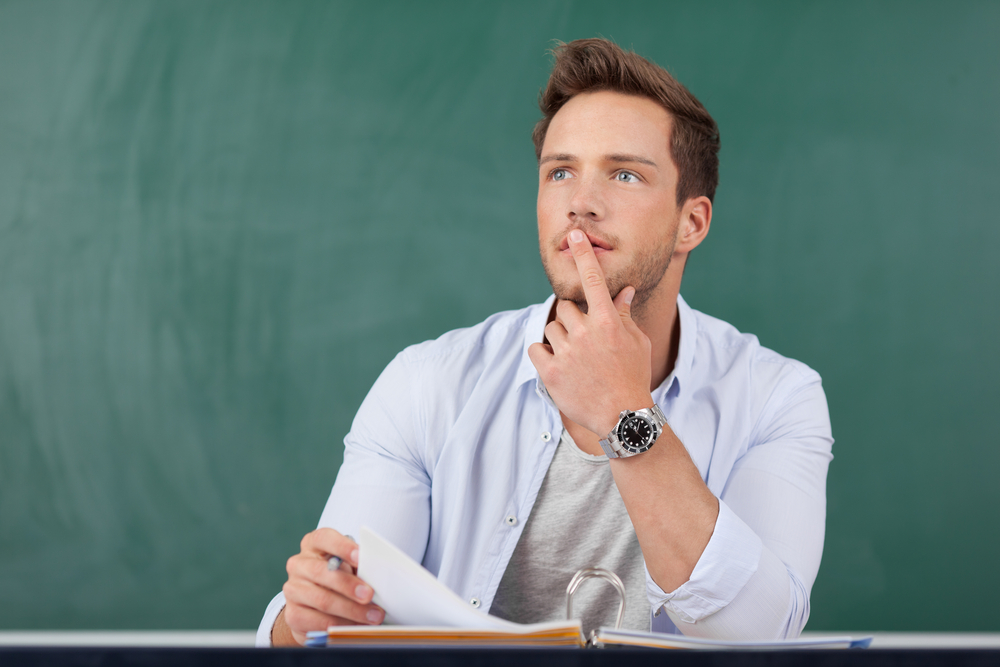 Thoughtful young man sitting with book in front of chalkboard